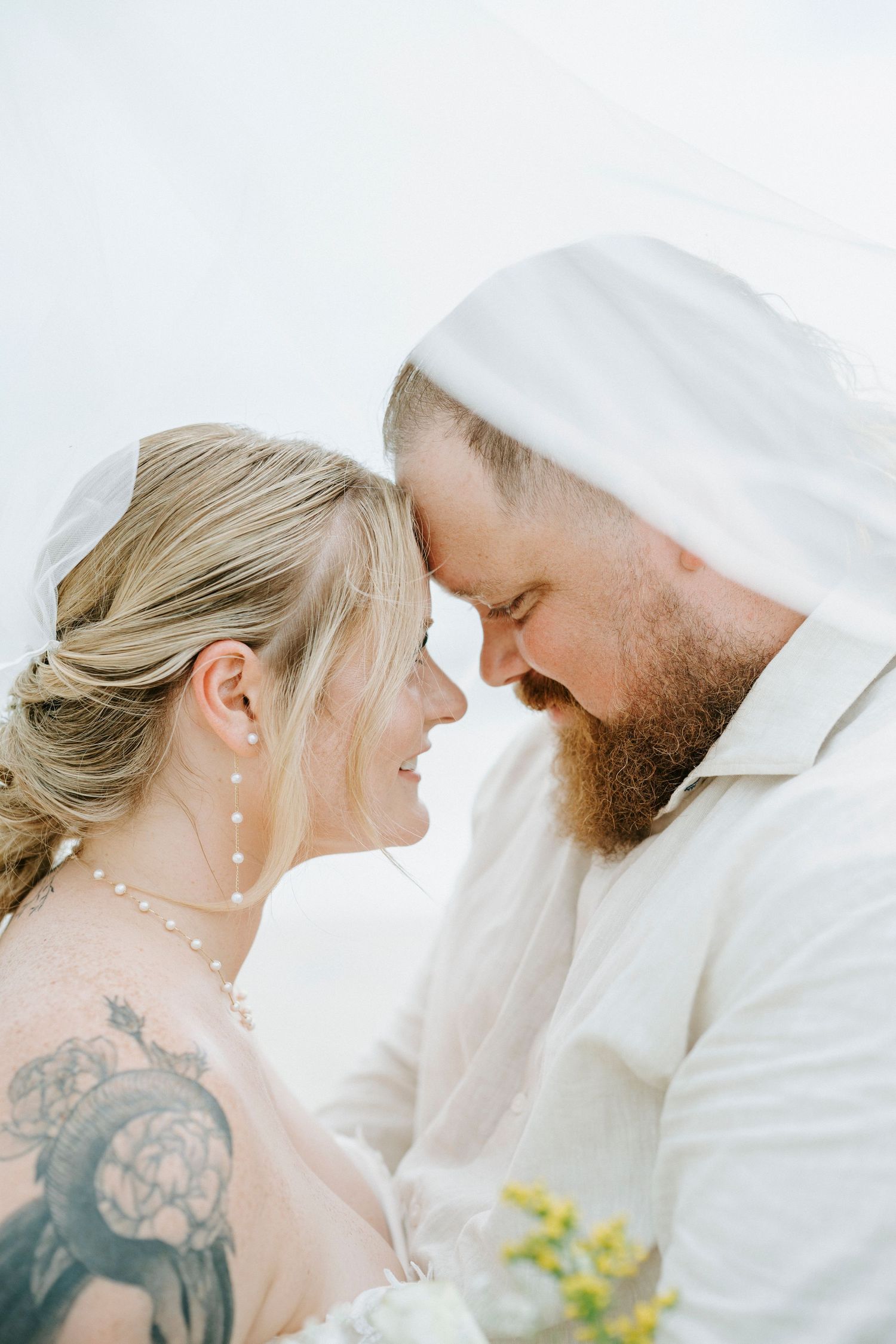 A romantic couple shares an intimate moment under a white veil against a bright background.
