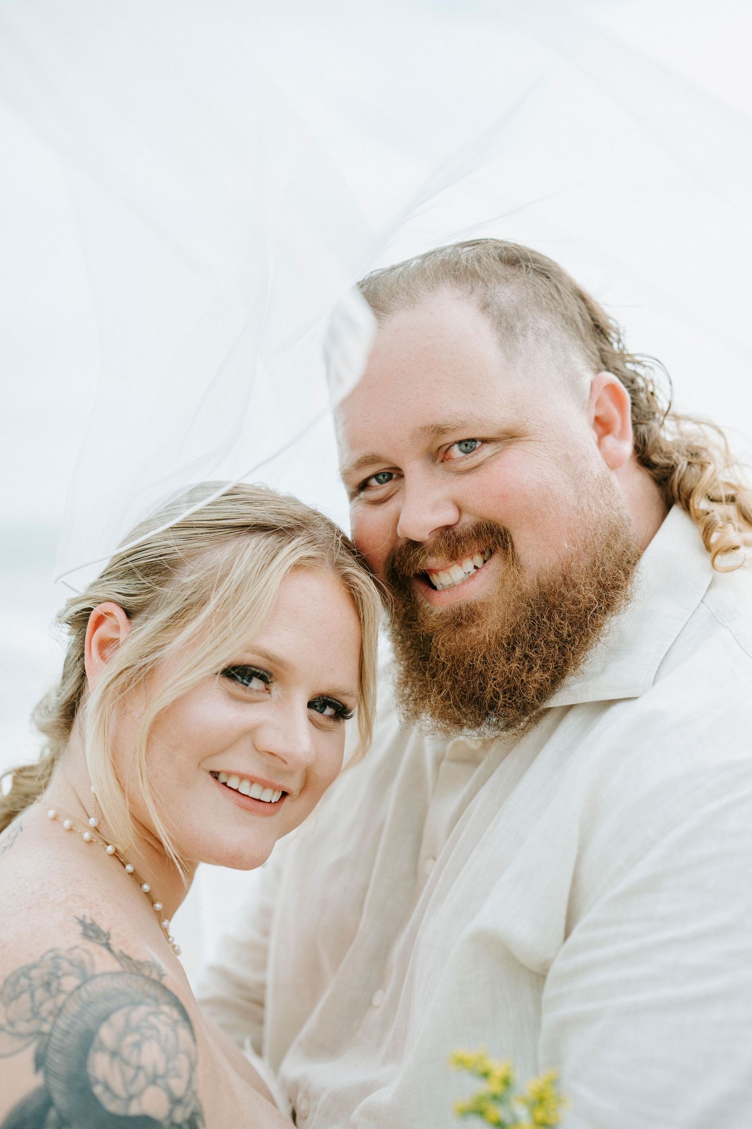 A happy couple embraces outdoors, with one person wearing white and showing visible tattoo artwork.