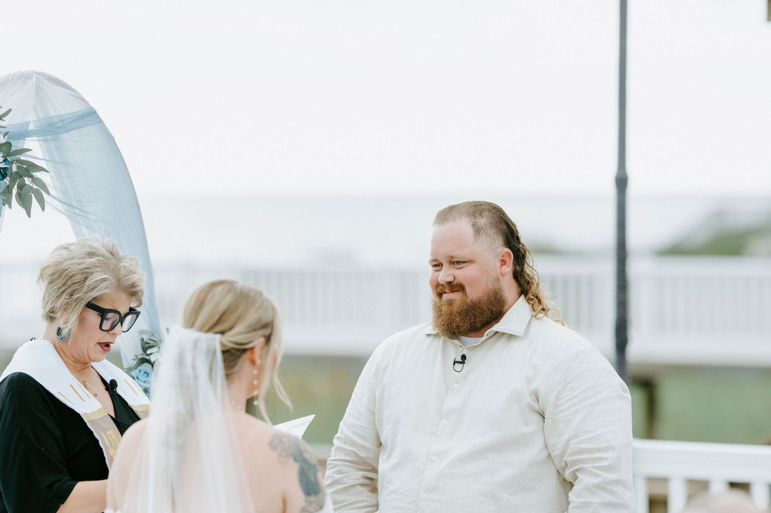 A couple stands together on an oceanfront balcony in coordinating cream-colored outfits with coastal views.