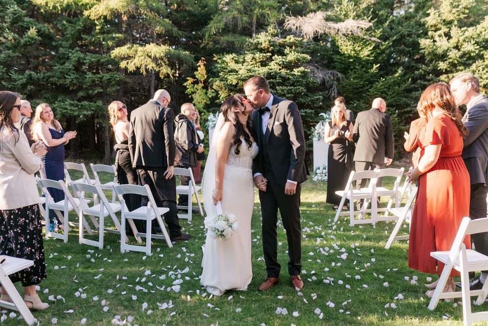 Wedding ceremony guests seated in white chairs on a garden lawn with scattered flower petals along the aisle.