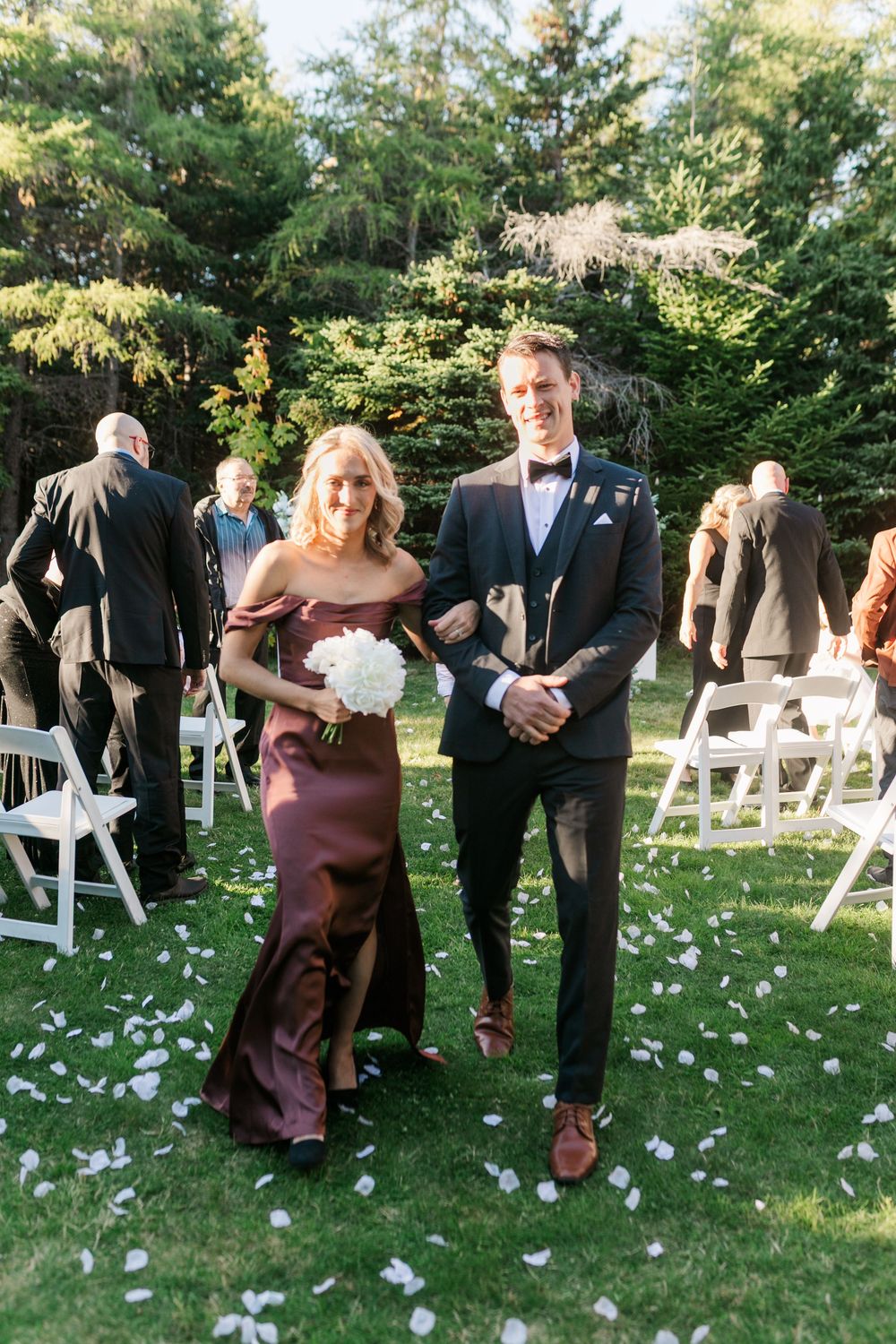 Wedding party members in formal attire stand in a line on grass with scattered white flower petals during an outdoor ceremony.