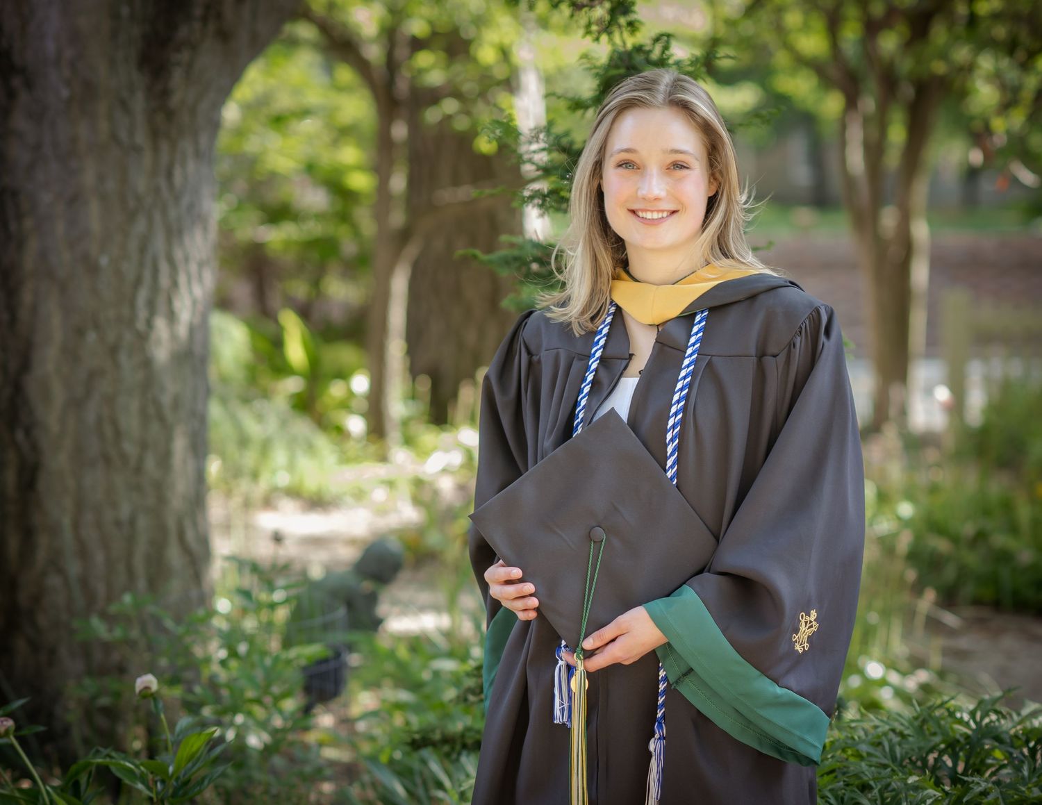 Smiling young woman in dark graduation gown with yellow and blue cords holding cap outdoors among trees Best photographer Williamsburg VA