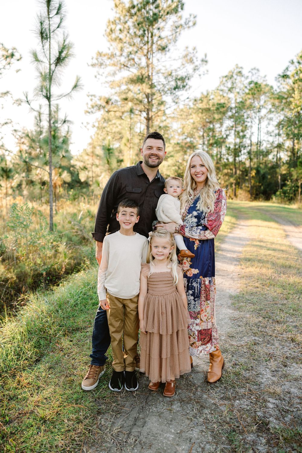 A family of five poses together on a wooded path during golden hour wearing coordinated casual outfits.