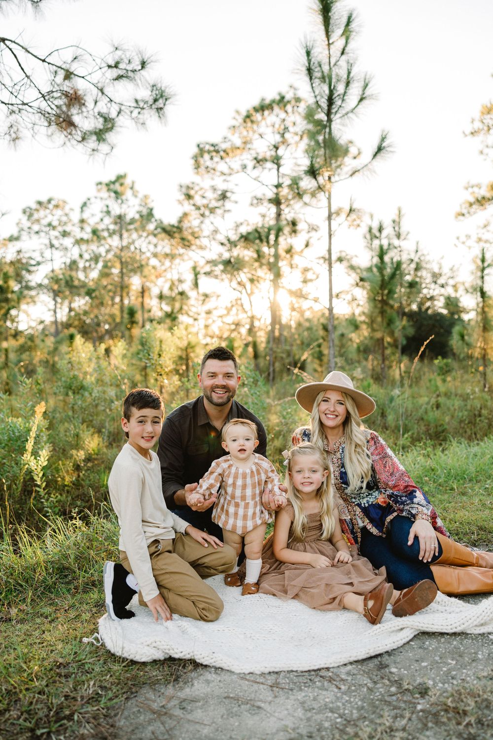 A family sits together on a white blanket outdoors surrounded by tall pine trees during a golden sunset.