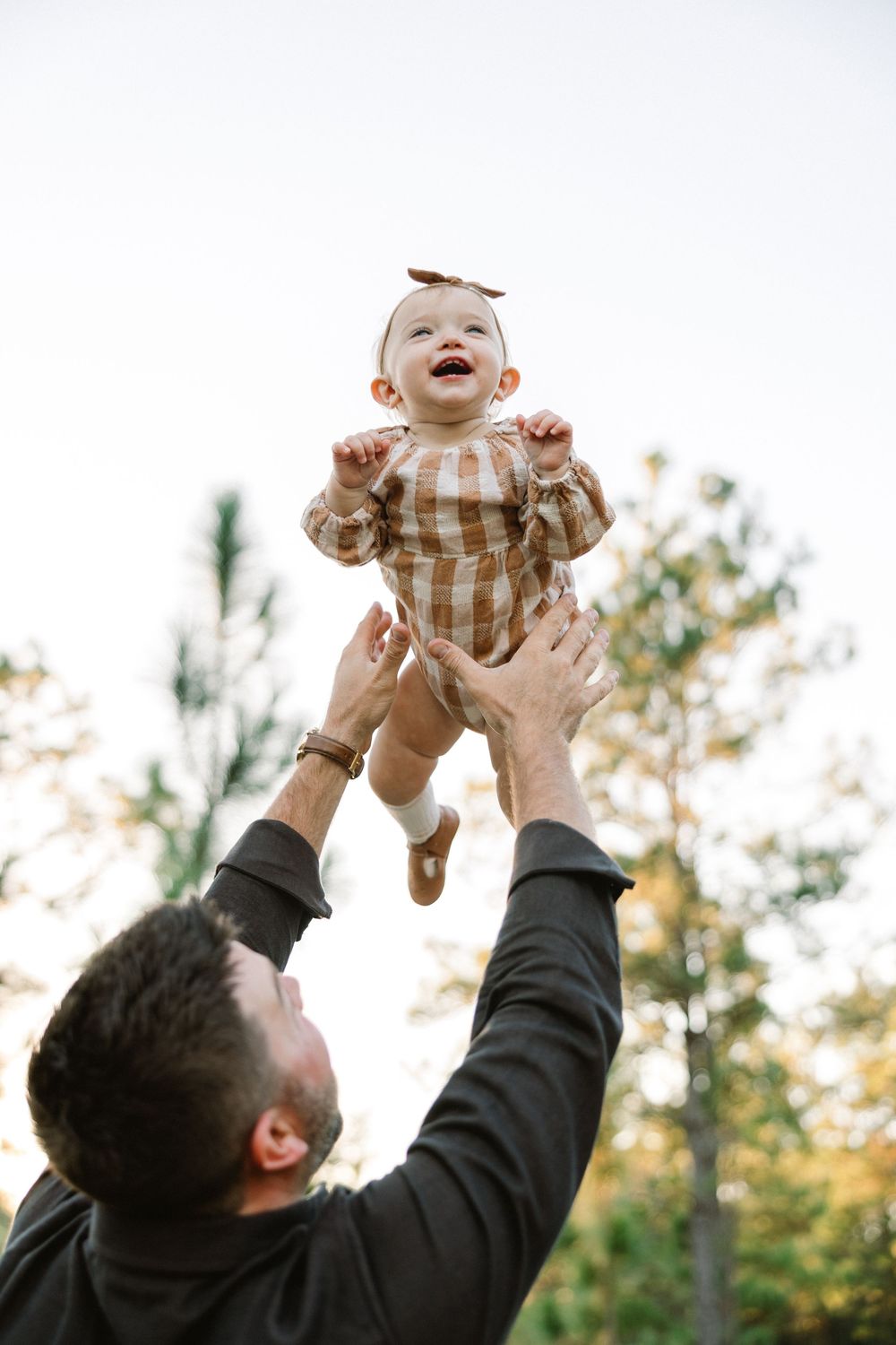 Parent playfully lifts baby in striped outfit up in the air against trees and sky at sunset.