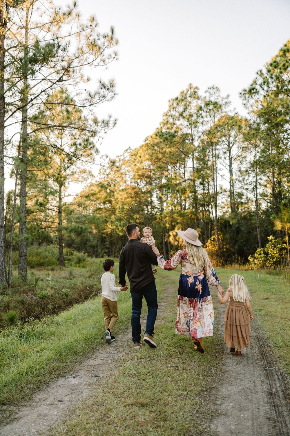 A group walks together down a scenic dirt path surrounded by tall pine trees at sunset.