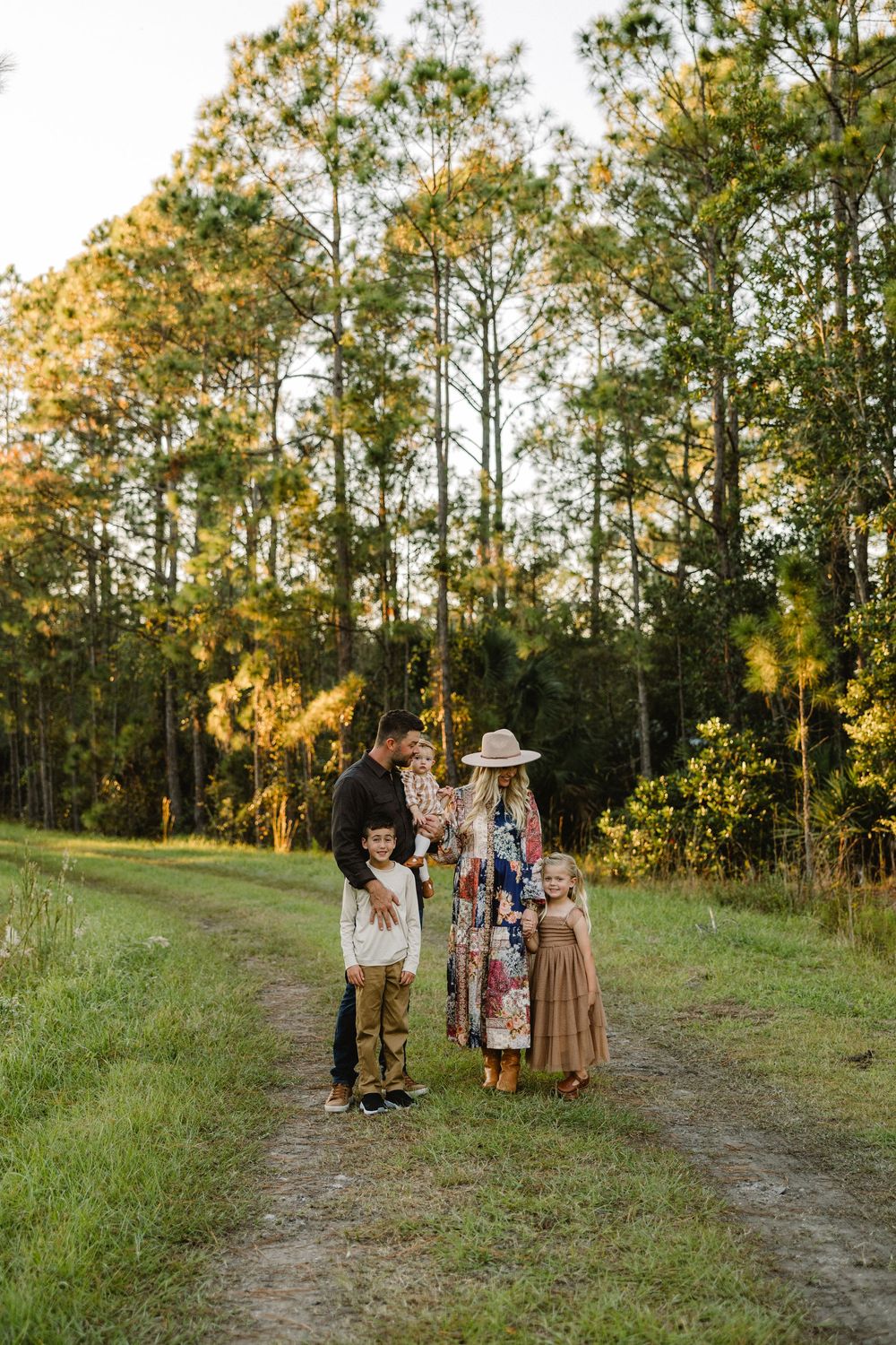 A group of people gather on a grassy trail at sunset with tall pine trees in the background.