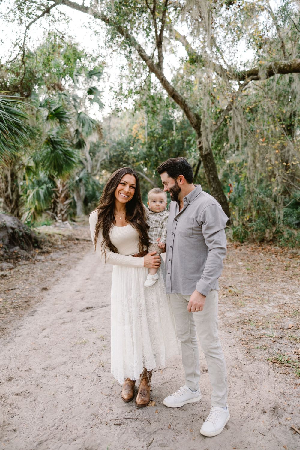 A couple and their baby walk along a sandy path surrounded by trees in a natural outdoor setting.