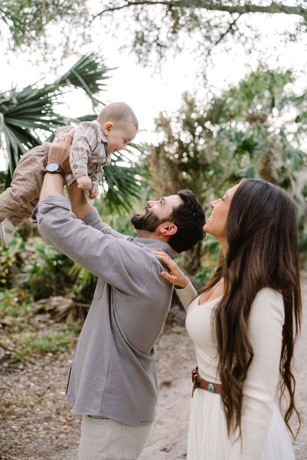 A couple shares a playful moment in nature, lifting their baby up together while smiling and laughing.