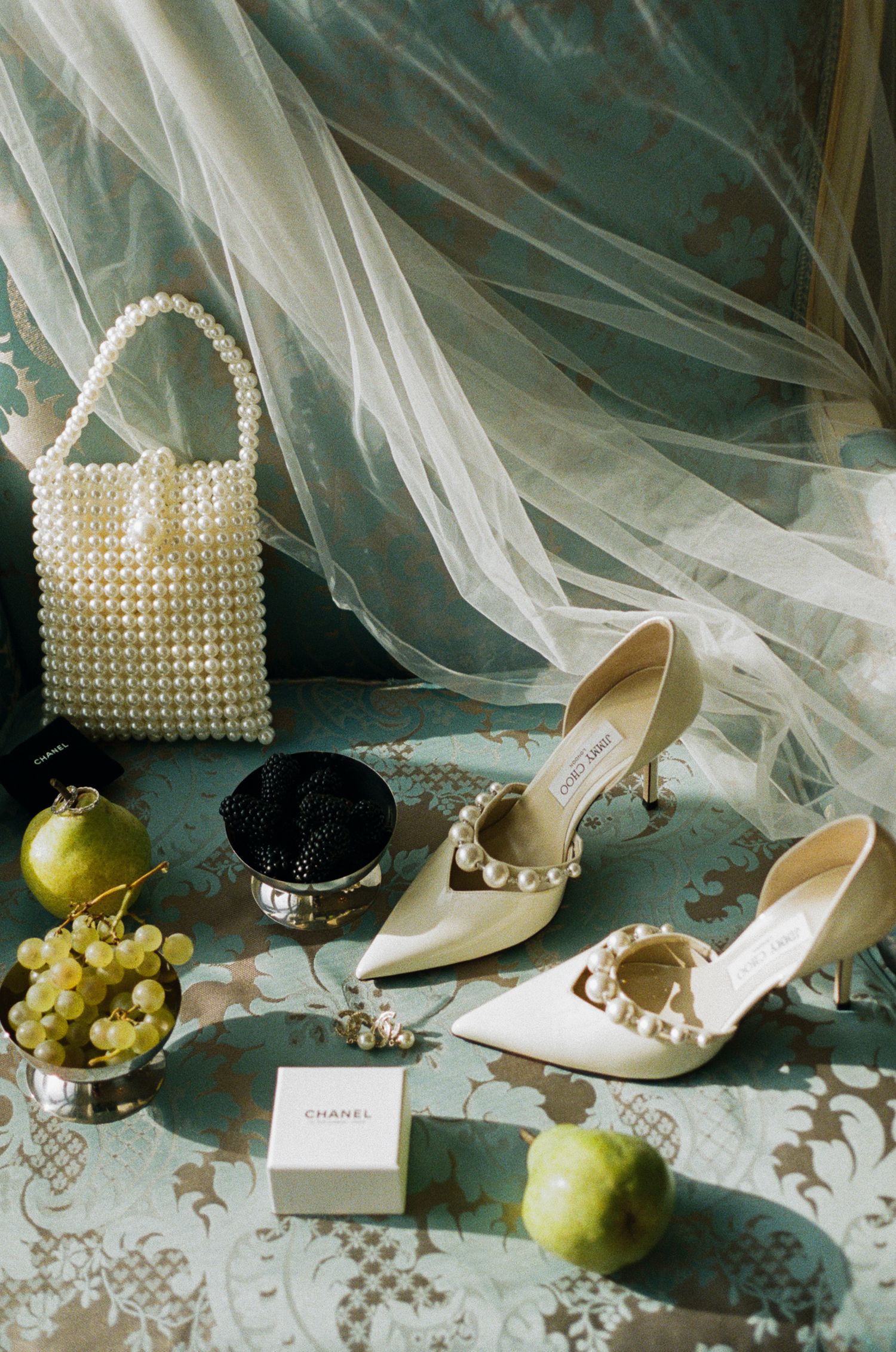Elegant bridal accessories including pearl purse, white heels, jewelry, and fresh grapes arranged on vintage lace fabric.
