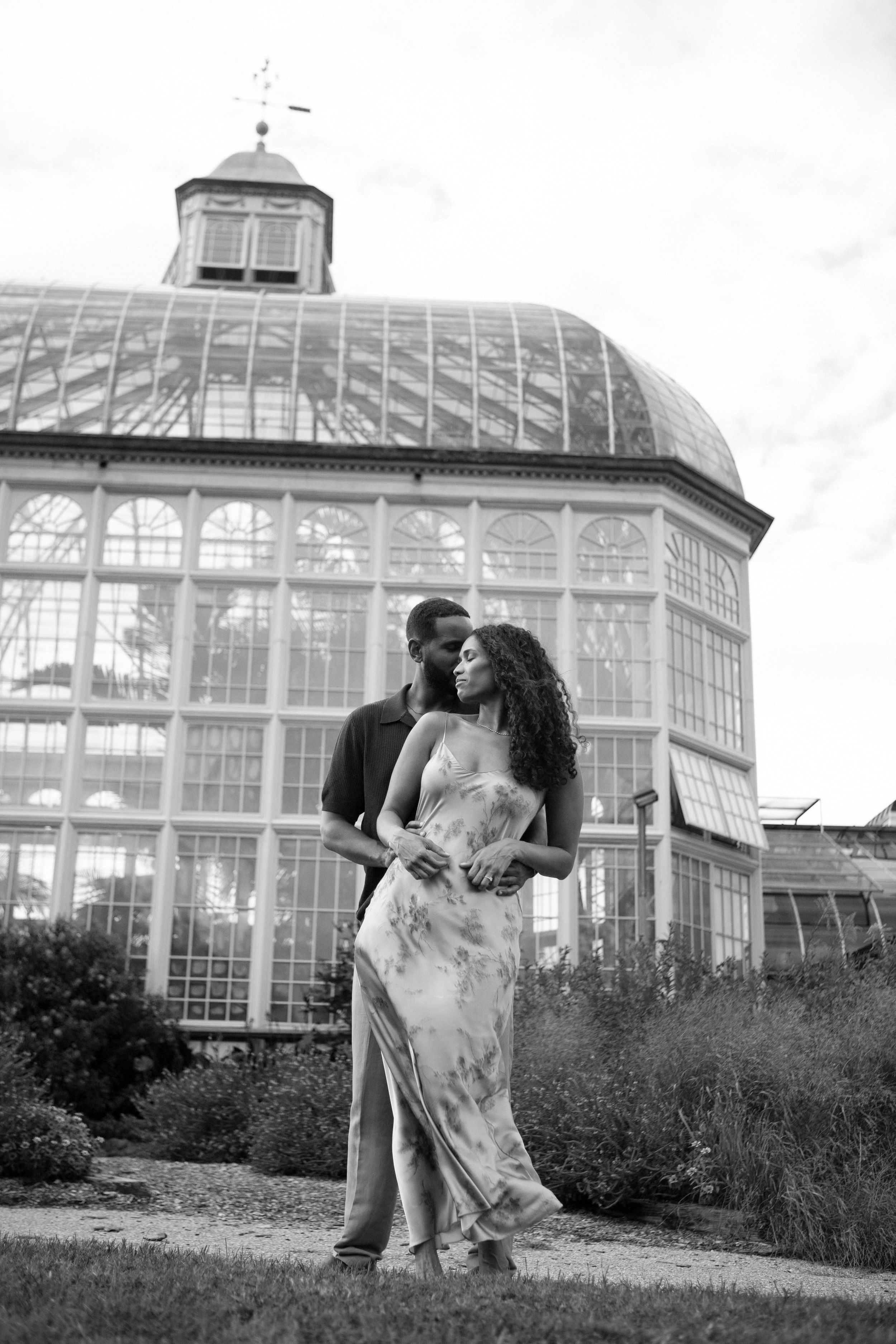 A romantic couple embraces in front of a glass conservatory building in a black and white photo series.