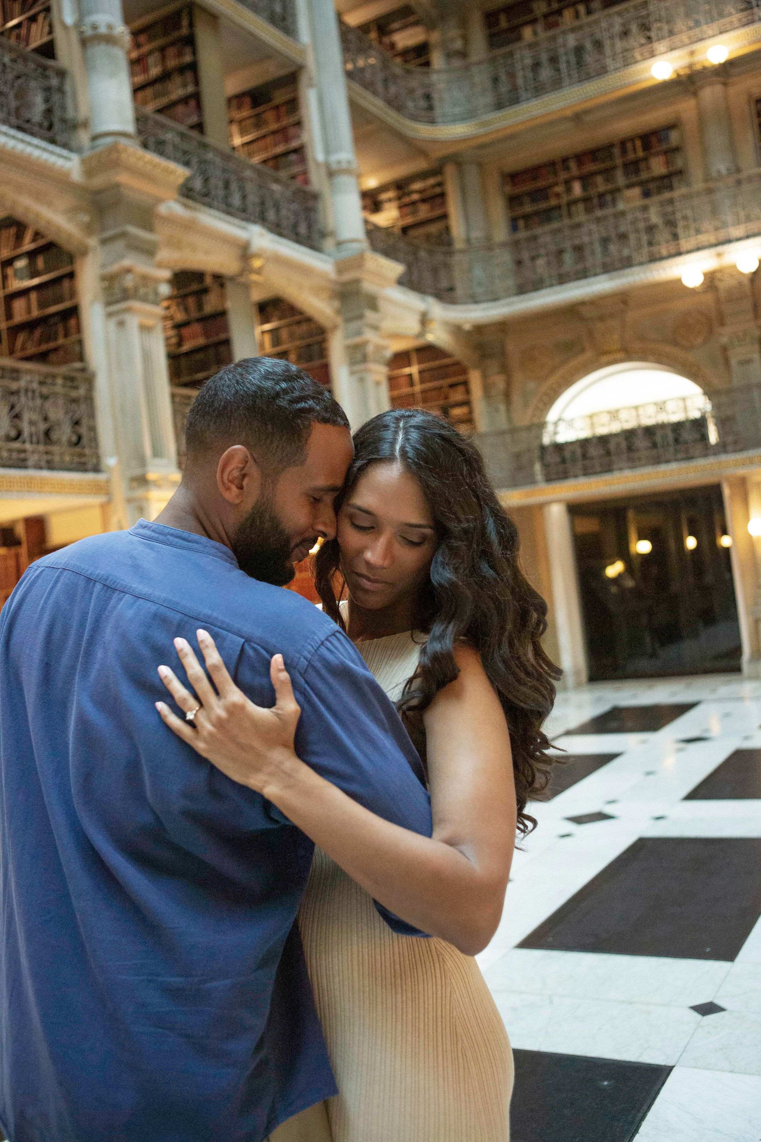 A couple shares an intimate embrace in an elegant library with towering bookshelves and marble columns in the background.