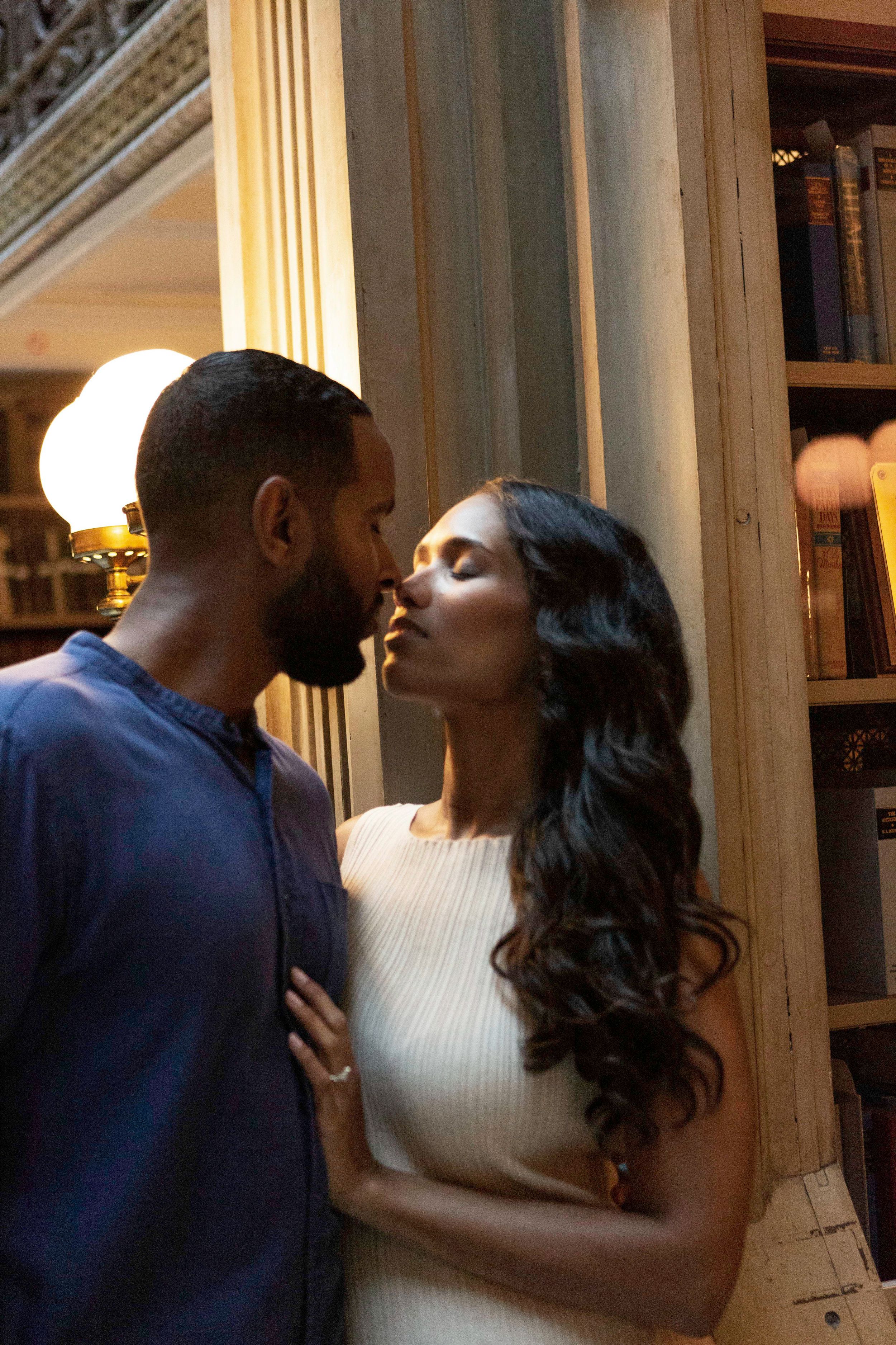 A romantic couple in formal attire share an intimate kiss against an elegant backdrop with bookshelves and columns.