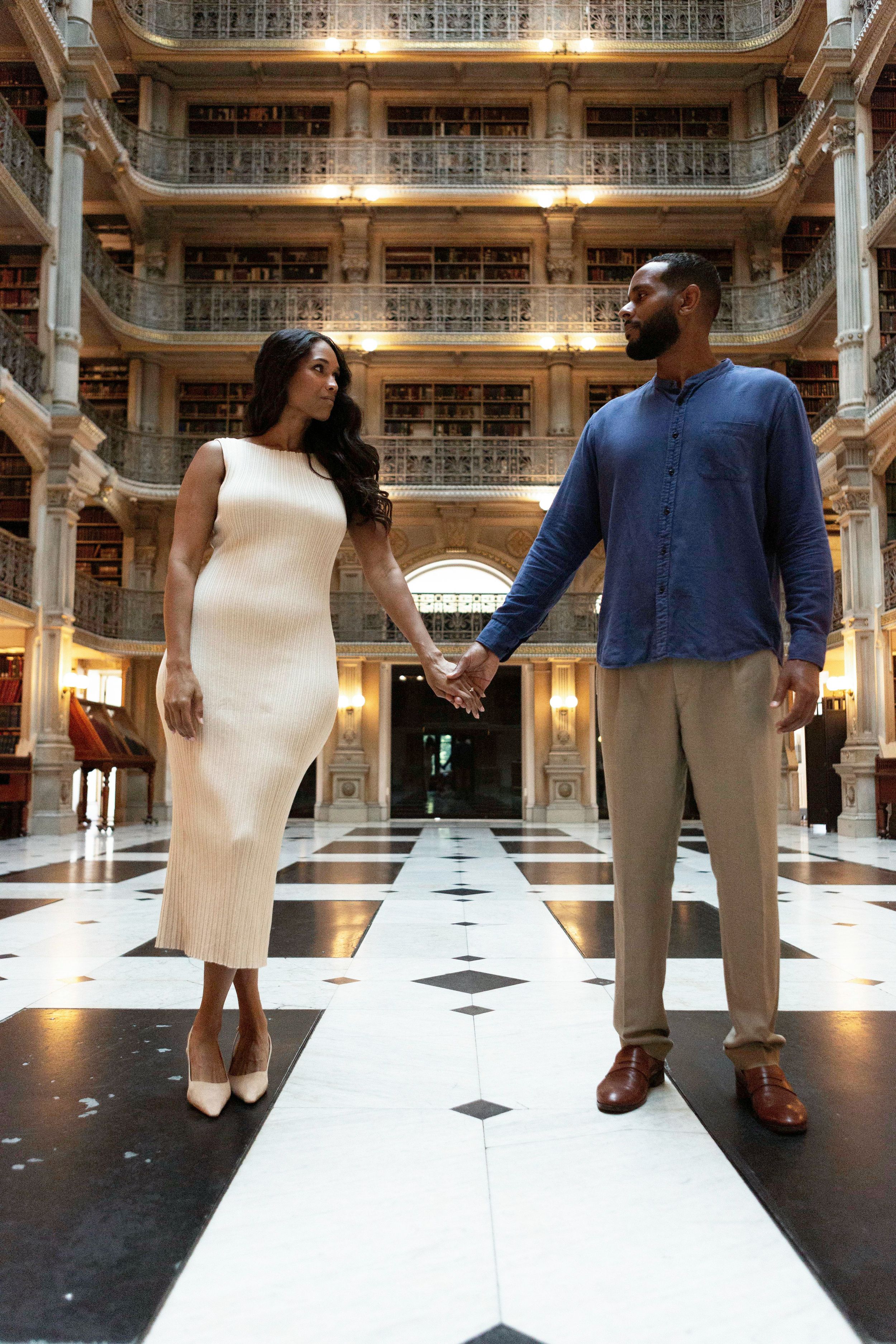 A couple holds hands while standing in an ornate historic library with multiple floors and elegant architecture.
