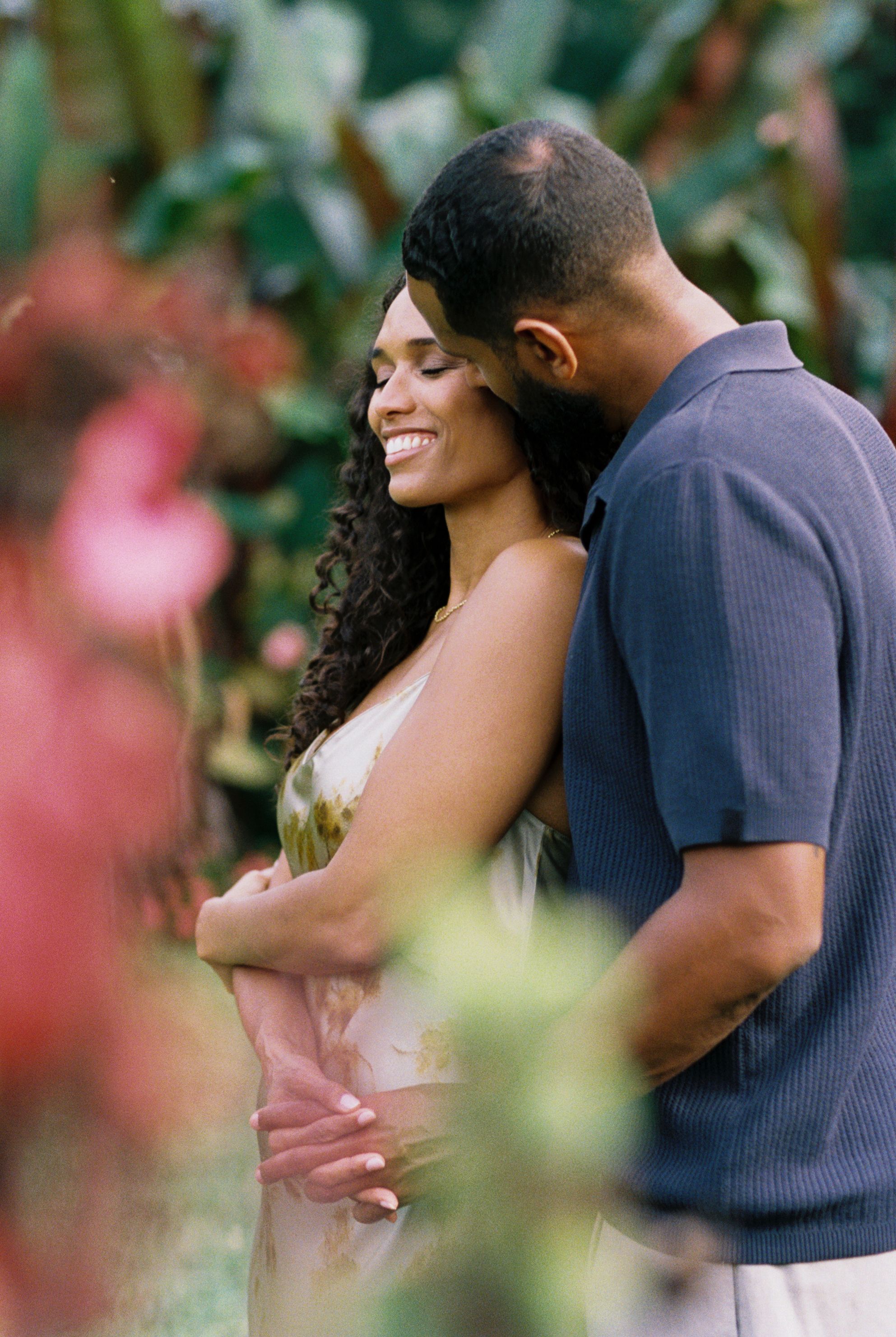 A romantic couple embraces and shares tender moments in a garden setting with soft pink floral bokeh in the background.