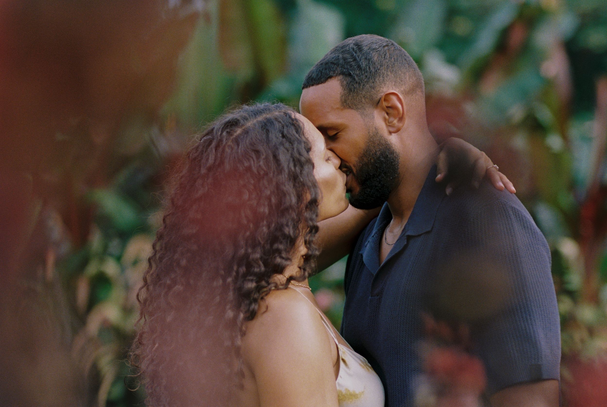 Romantic couple embraces in a lush garden setting with soft natural lighting and greenery in the background.