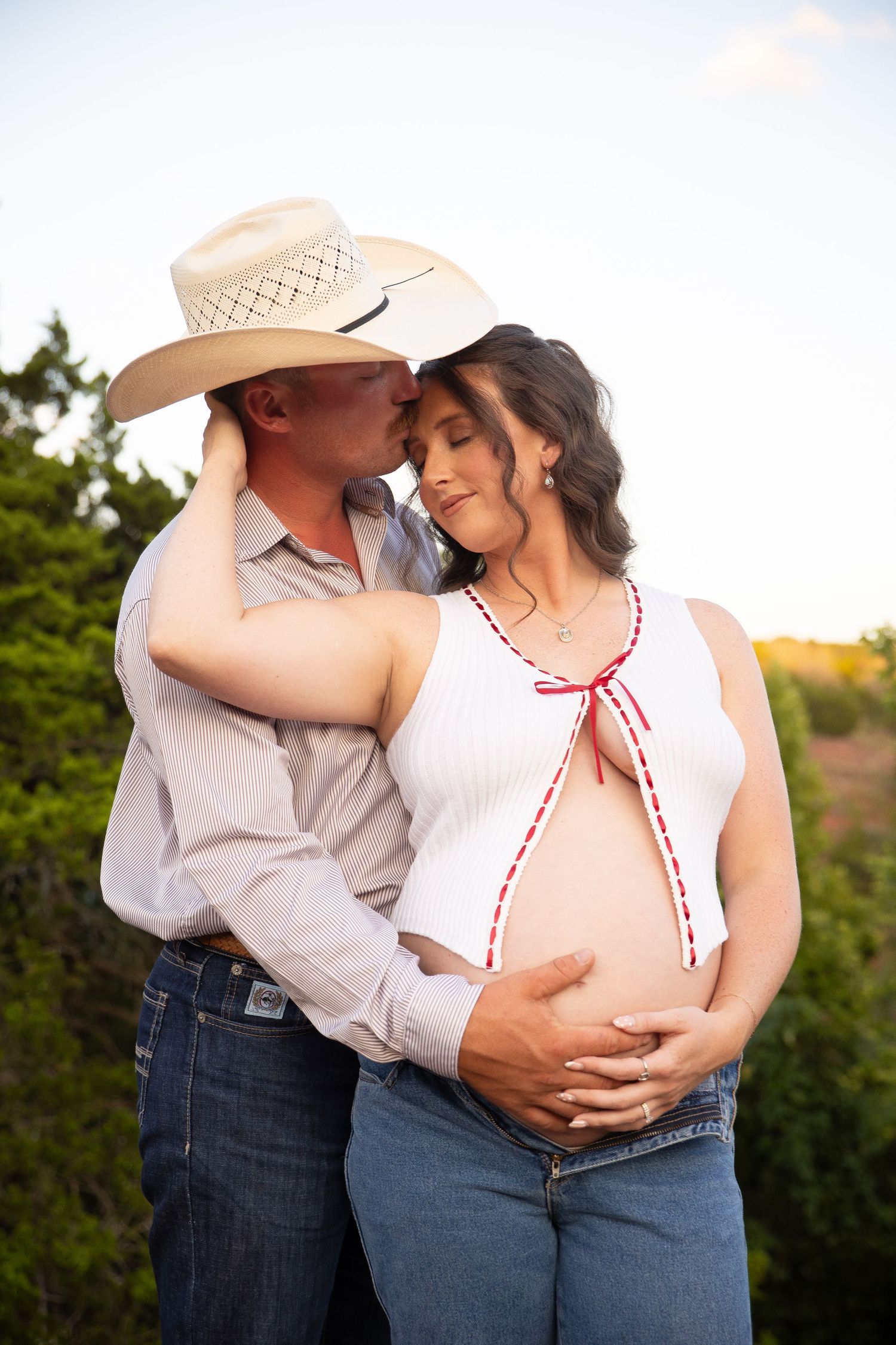 A romantic maternity photo series showing a couple embracing in casual western attire during sunset outdoors.