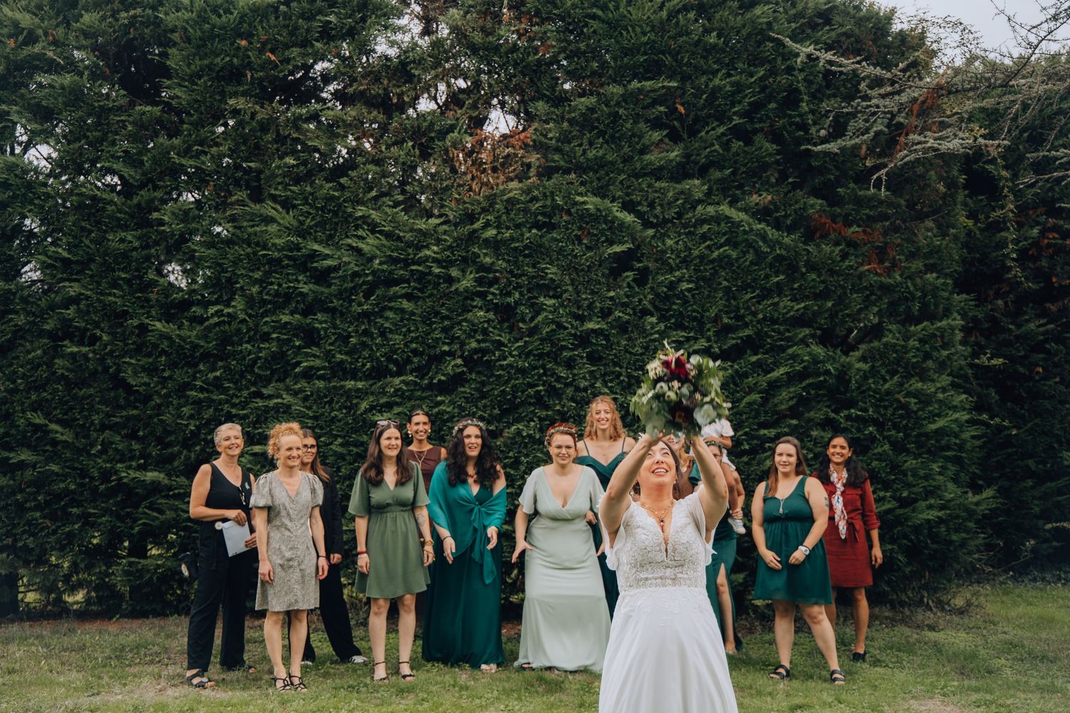 Bride tossing bouquet to group of women in green dresses at outdoor wedding ceremony.