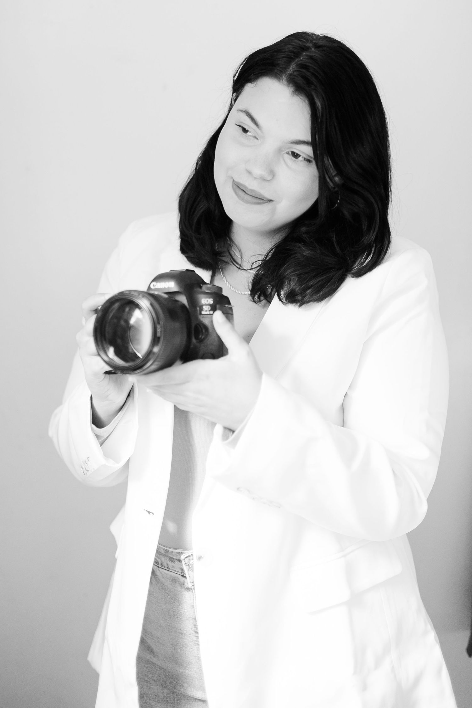 Professional photographer in white blazer holds DSLR camera while posing against light background in black and white portrait morgantown wv