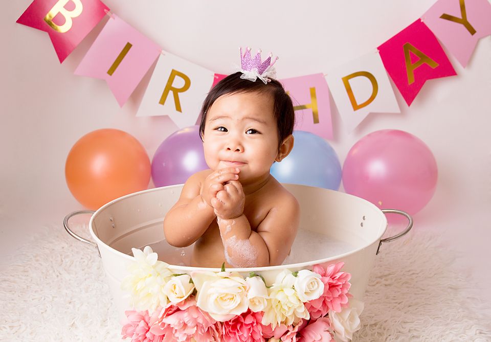 Baby in pink floral decorated bathtub with birthday banner and balloons in background.