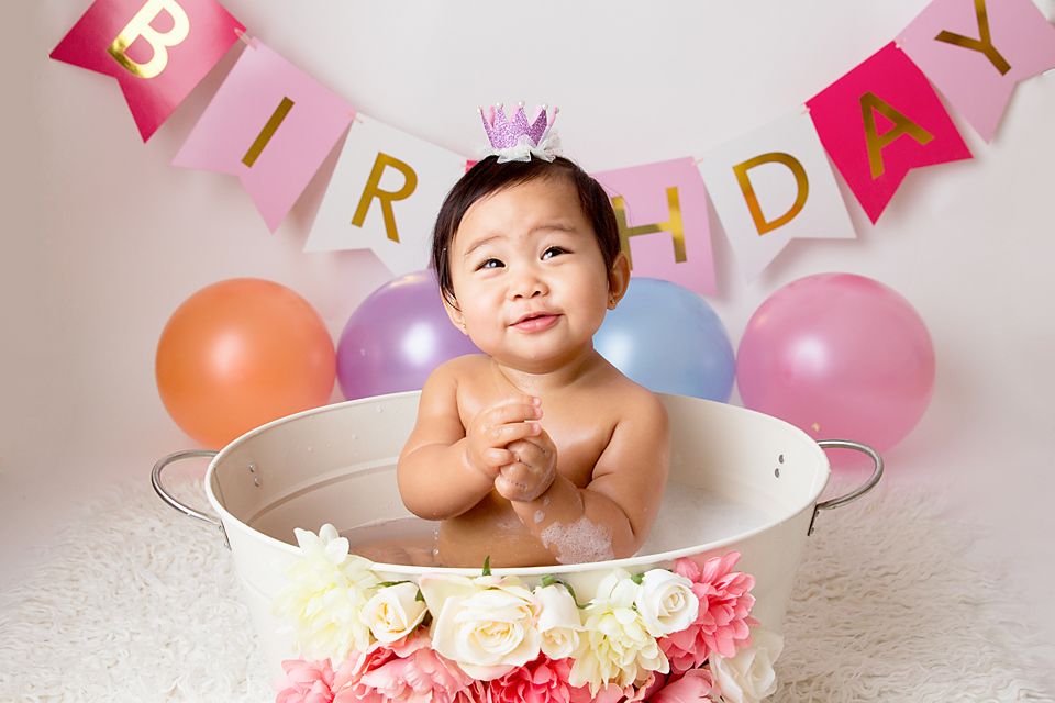 Baby posing in decorated bathtub with pink and gold birthday banner and pastel balloons.