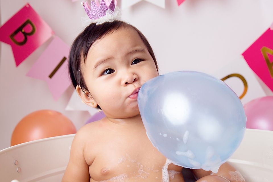 Baby lying in white bathtub surrounded by pink and gold birthday decorations.