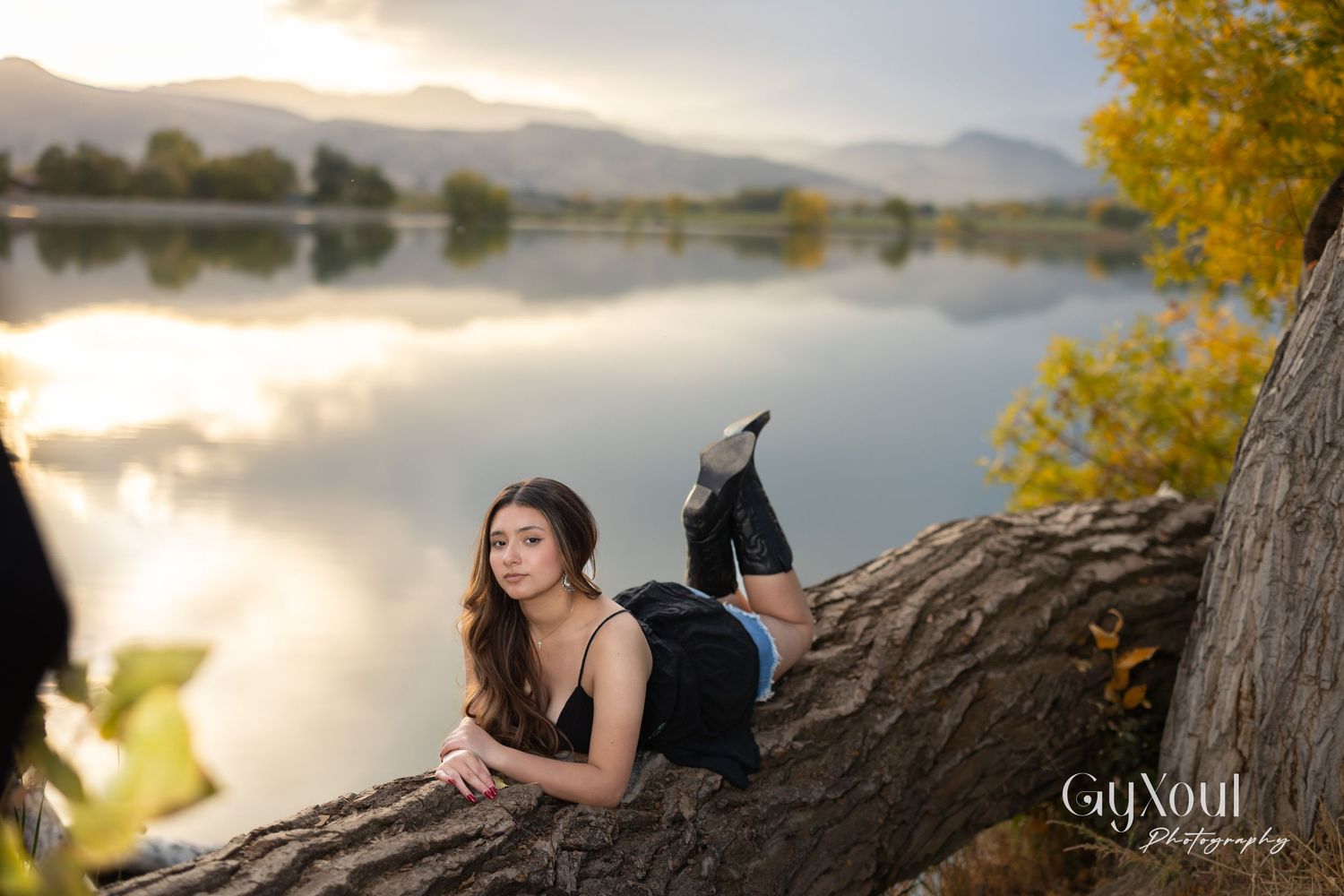 Person lying on fallen tree trunk by tranquil lake during sunset with mountain backdrop and fall foliage.