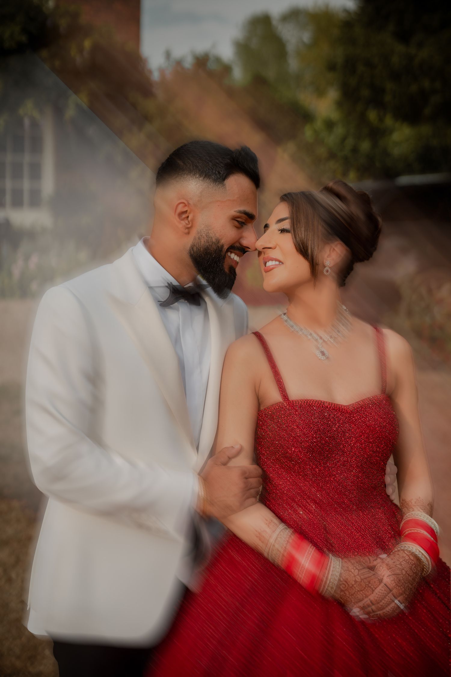 A romantic couple in formal attire, with the woman in a red gown and the man in white, share intimate moments outdoors.