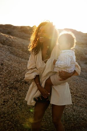 A parent and child share a tender moment at sunset on rocky terrain, backlit by golden sunlight.