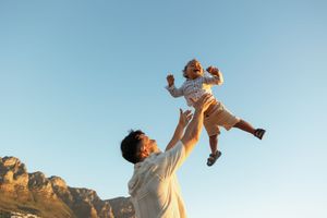 A parent lifts a joyful toddler high into the air against a backdrop of mountains and blue sky at sunset.