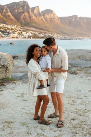 A family shares tender moments together on a rocky beach with scenic mountain cliffs in the background at sunset.