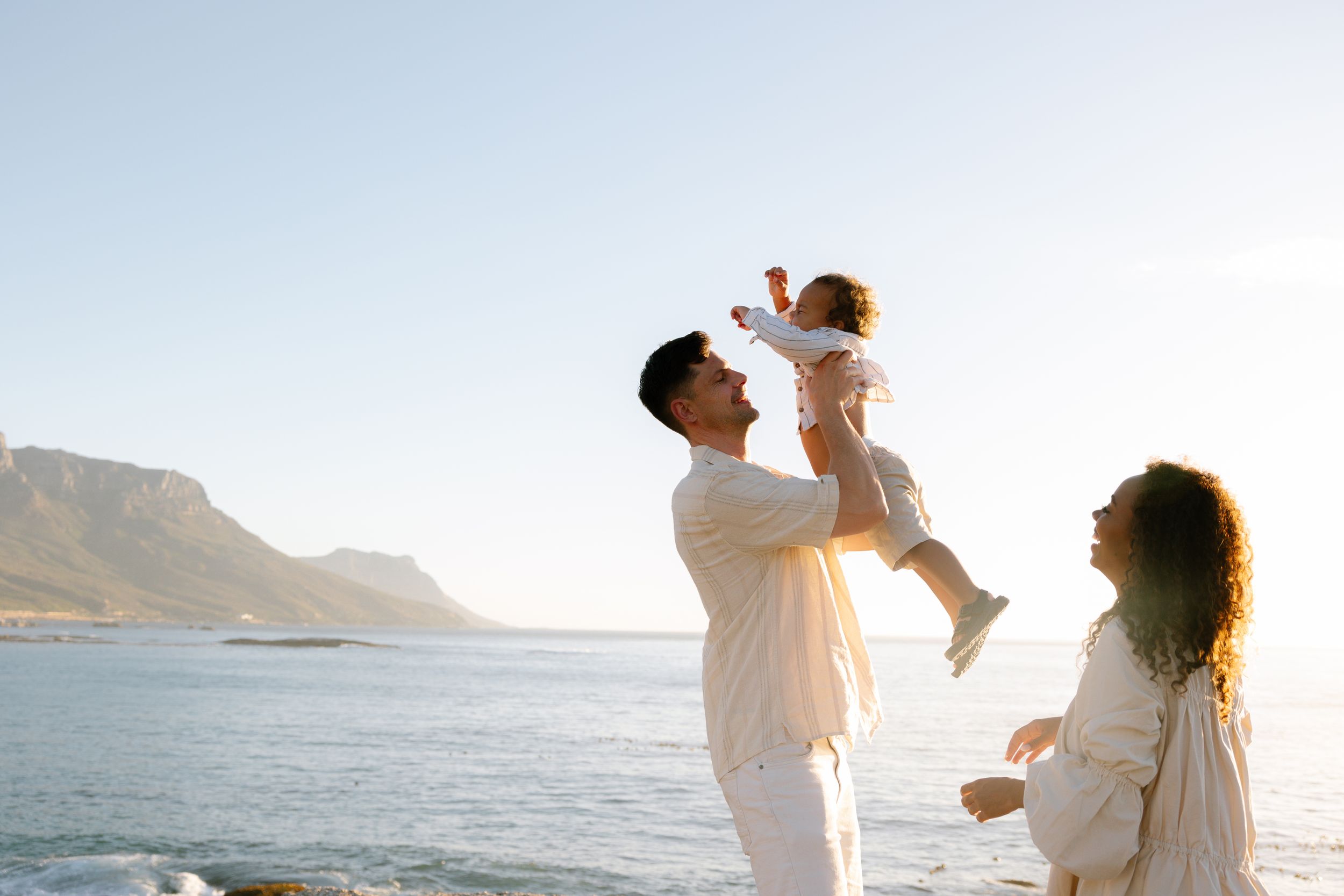 A couple in white outfits shares a romantic moment on a beach during sunset with mountains in the background.