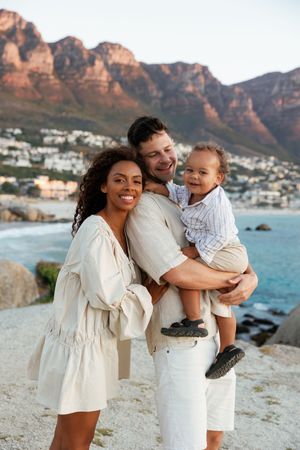 A family in white outfits poses together on a scenic coastal cliff with mountains and ocean views in the background.