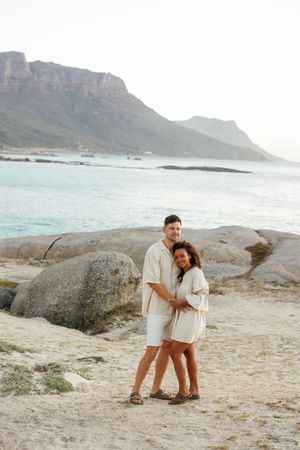 A couple shares tender moments on a scenic beach with dramatic mountain cliffs and ocean views in the background.