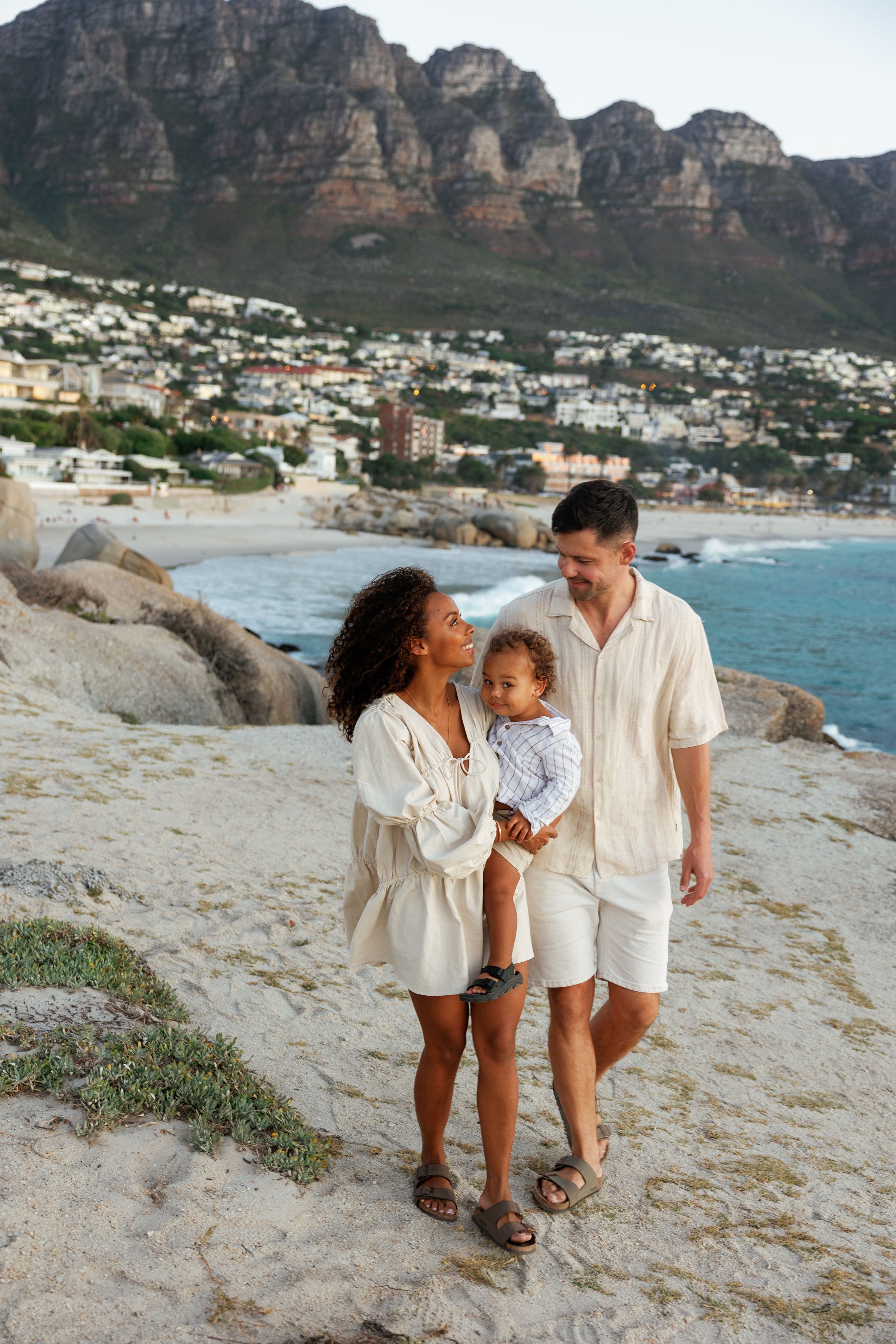 A family enjoys a tender moment together on a scenic beach with mountains and coastal homes in the background.