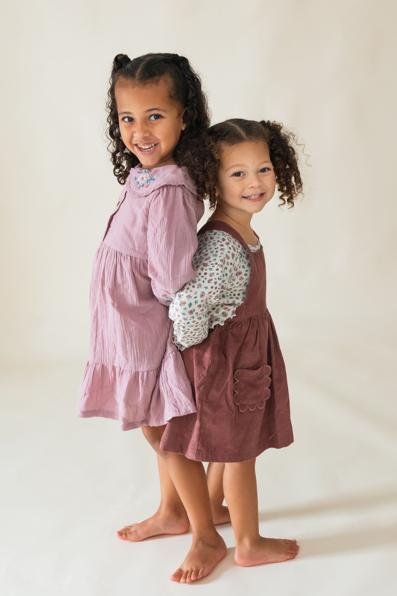 Two children in tiered dresses stand back-to-back and smile while posing barefoot against a light backdrop.