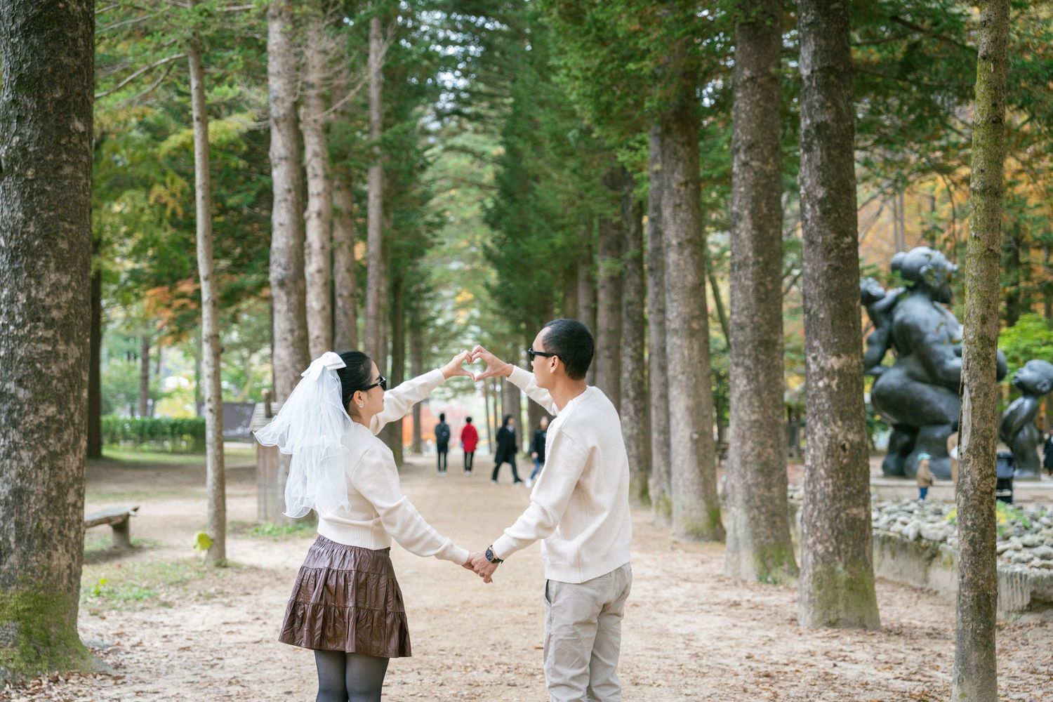 A couple holds hands while walking through a scenic tree-lined path in a park on a sunny day.