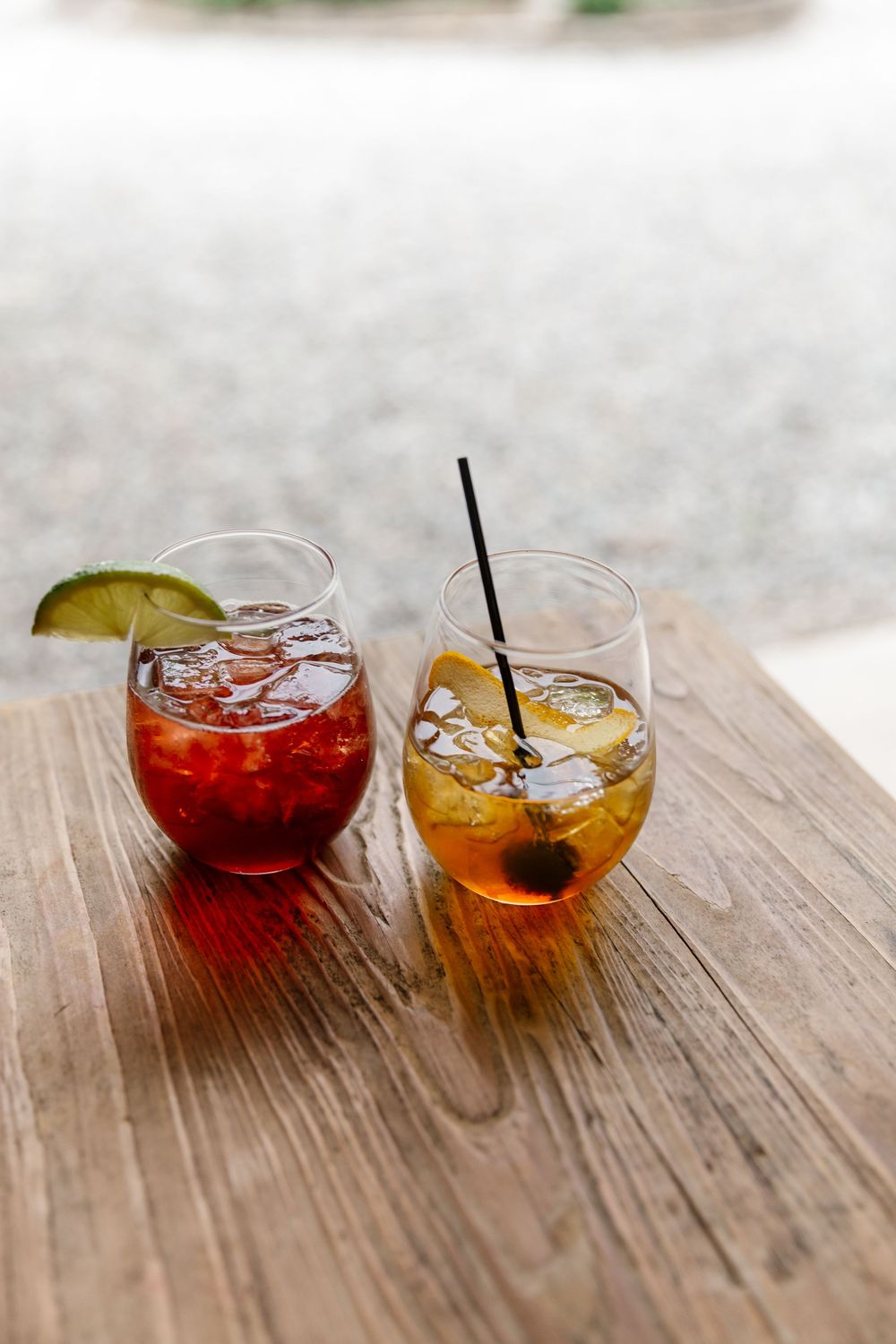 Two cocktails with ice and garnish on wooden table, one red and one amber colored.
