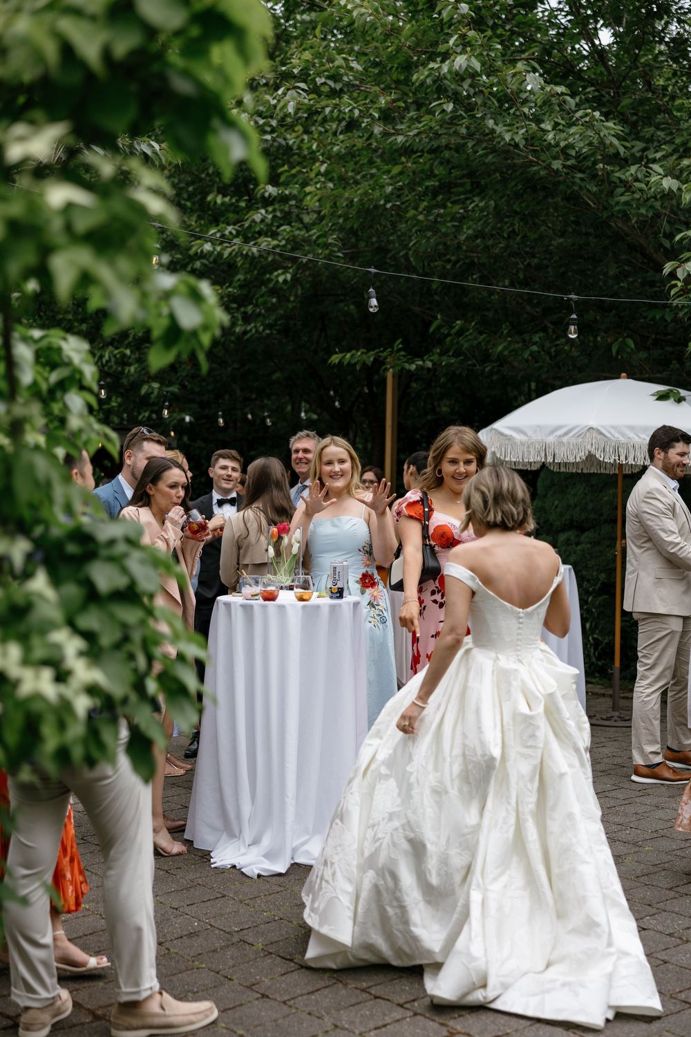 Wedding reception setup outdoors with white-clothed tables and guests mingling under lush green trees.