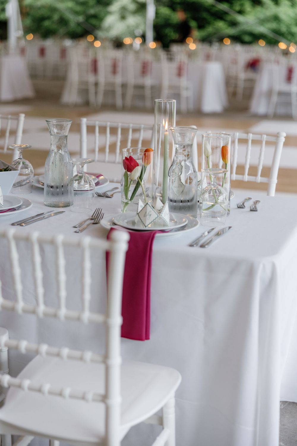 Elegant wedding table setting with white linens, chiavari chairs, glass vases, and burgundy napkins at an outdoor reception.
