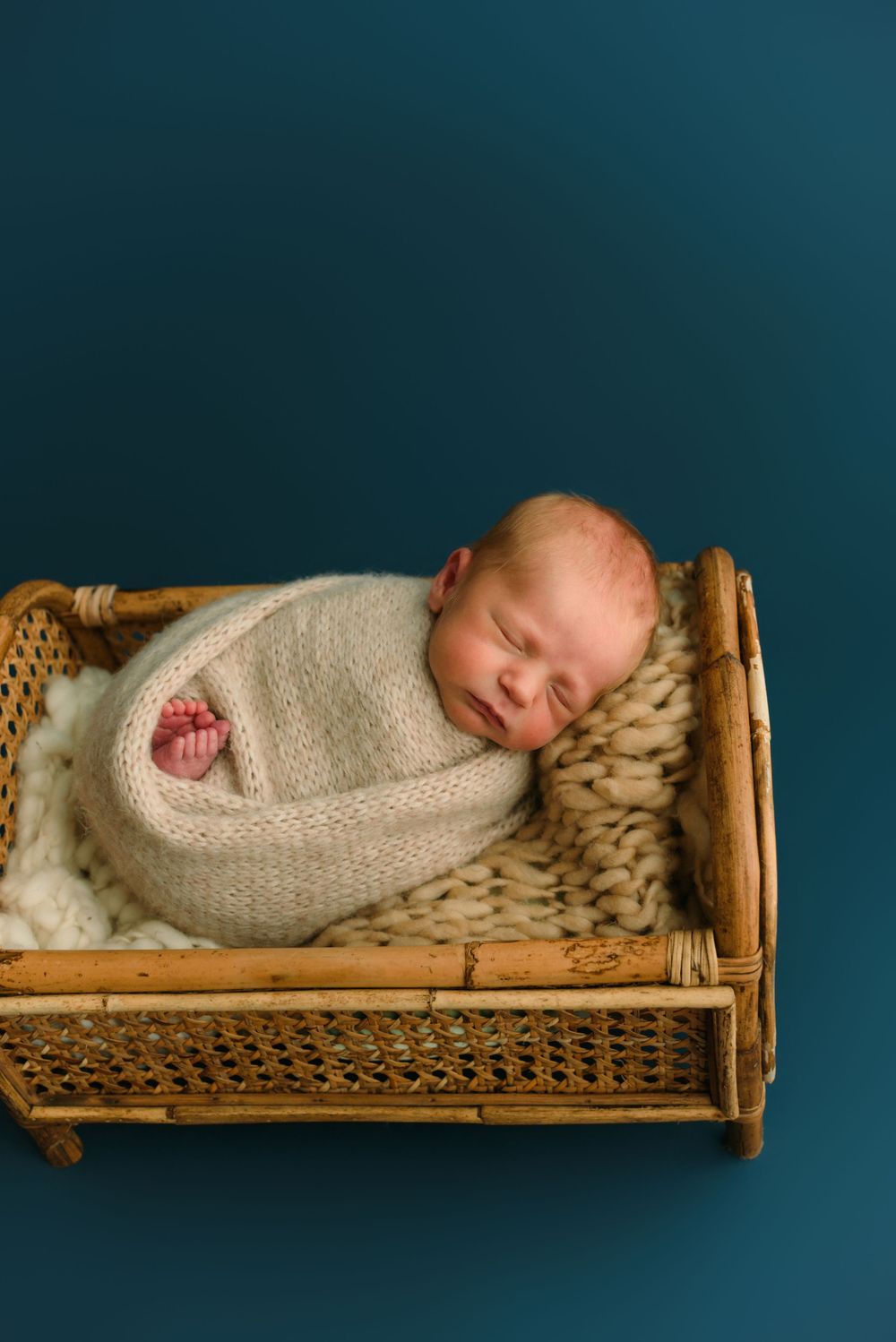 Newborn sleeping in knit wrap on vintage wicker bassinet.