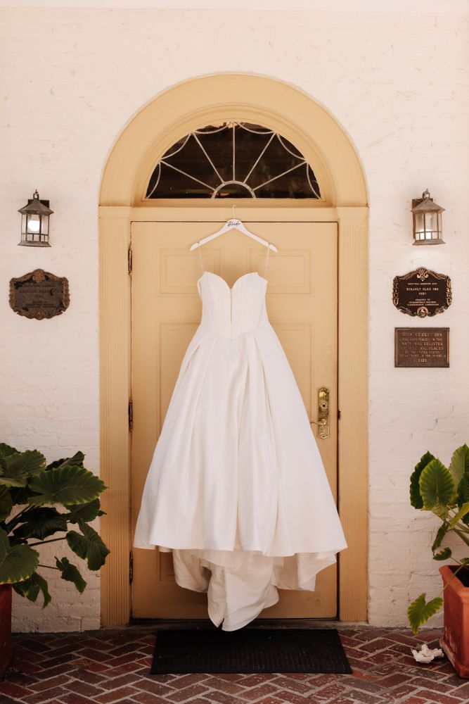 A white strapless wedding dress hangs on a hanger against a cream-colored arched doorway with lanterns on either side.