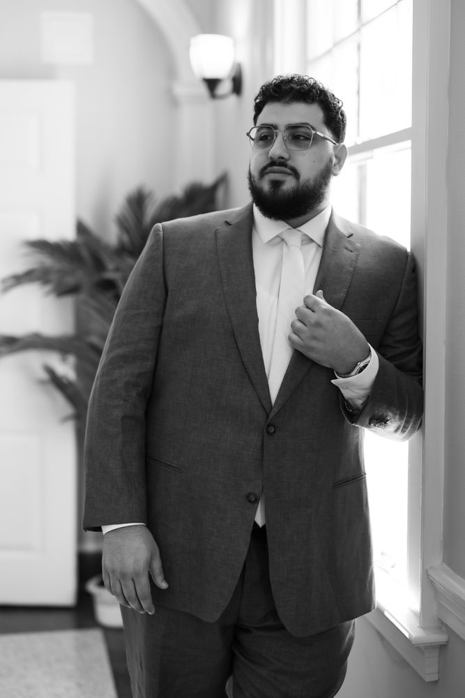 A groom in a dark suit and tie adjusts his collar while standing in a bright indoor setting.