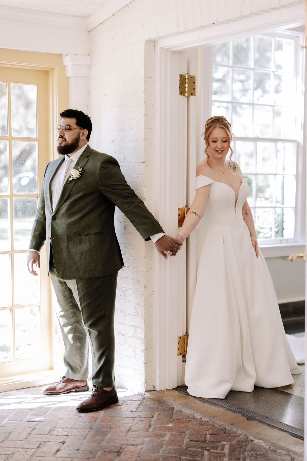 A couple shares a playful first look moment around a doorway corner in their wedding attire.