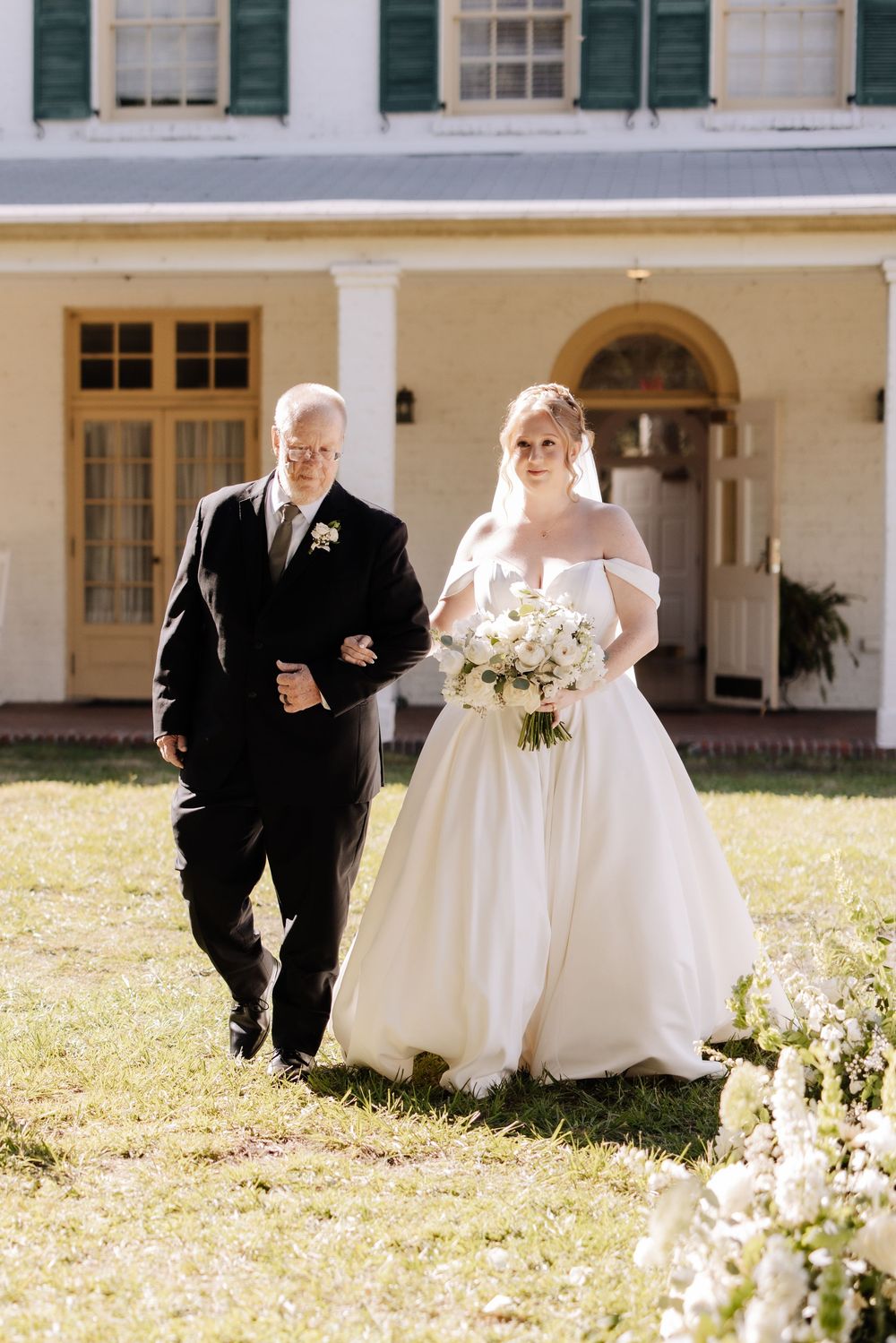 Wedding party members walk in a line across a sunlit lawn in front of a white colonial-style building.