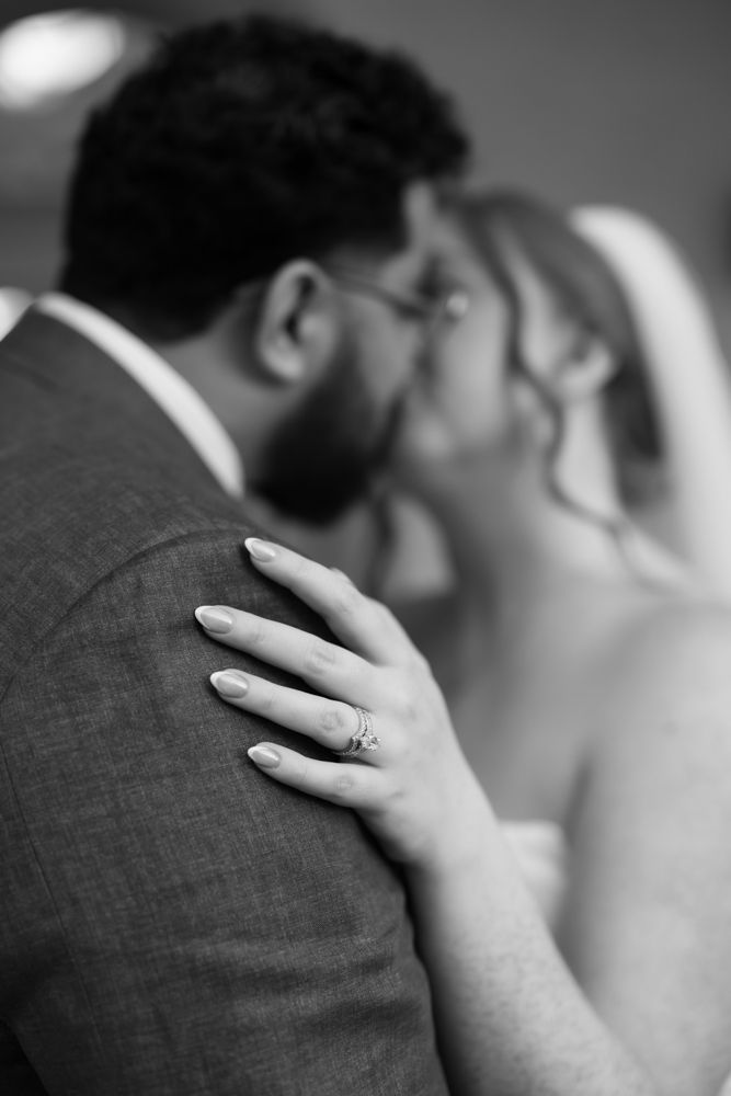Black and white close-up photo showing a hand with engagement ring resting on a suit jacket shoulder.