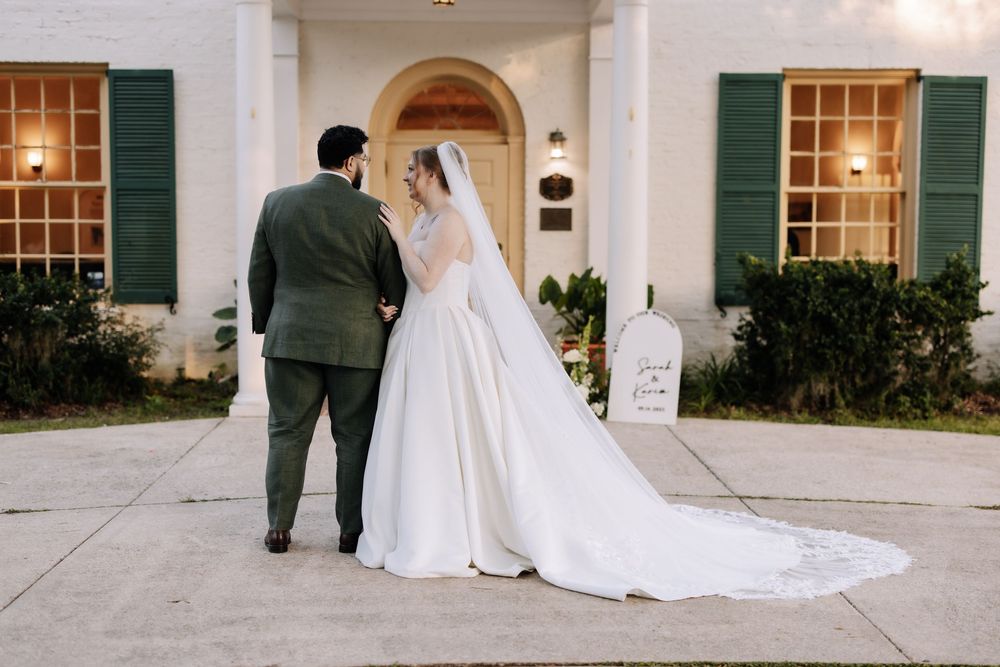 Elegant wedding couple poses in front of a white colonial style mansion with green shutters and arched windows.