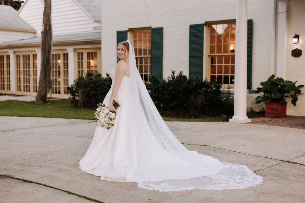 A bride in a flowing white wedding gown with long cathedral veil stands outside a white building with green shutters.