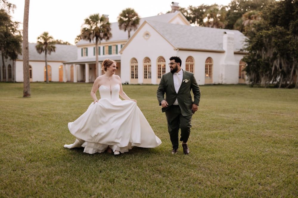 Wedding couple walks hand in hand on a grassy lawn with palm trees and a white church in the background.