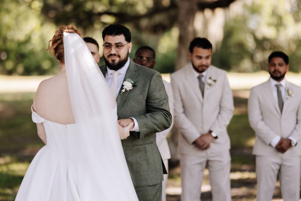 A bride and groom share their first look moment in a sunlit garden surrounded by wedding party members.